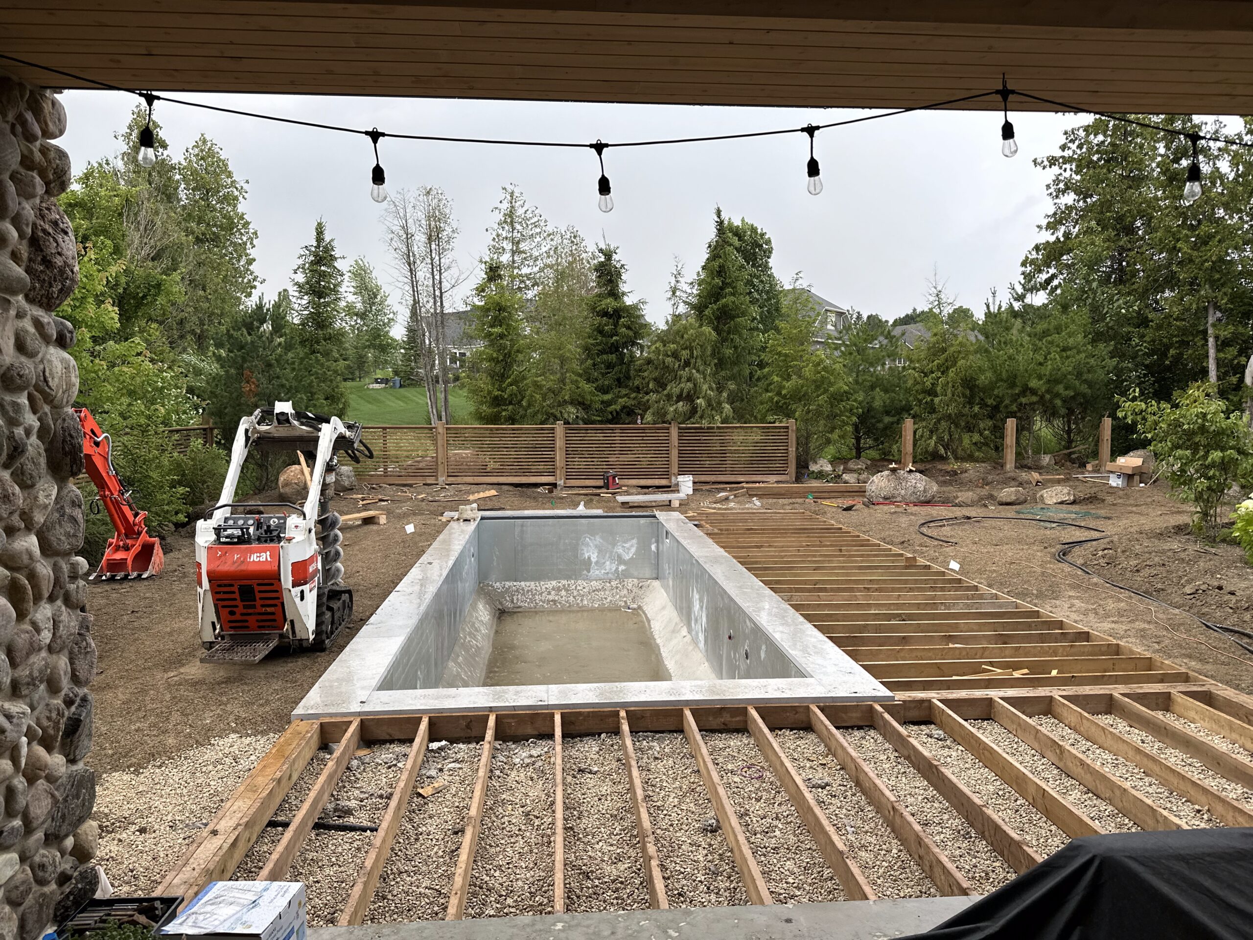 swimming pool area under construction, inground pool shell with deck beams surrounding it in dirt with small backhoe beside pool