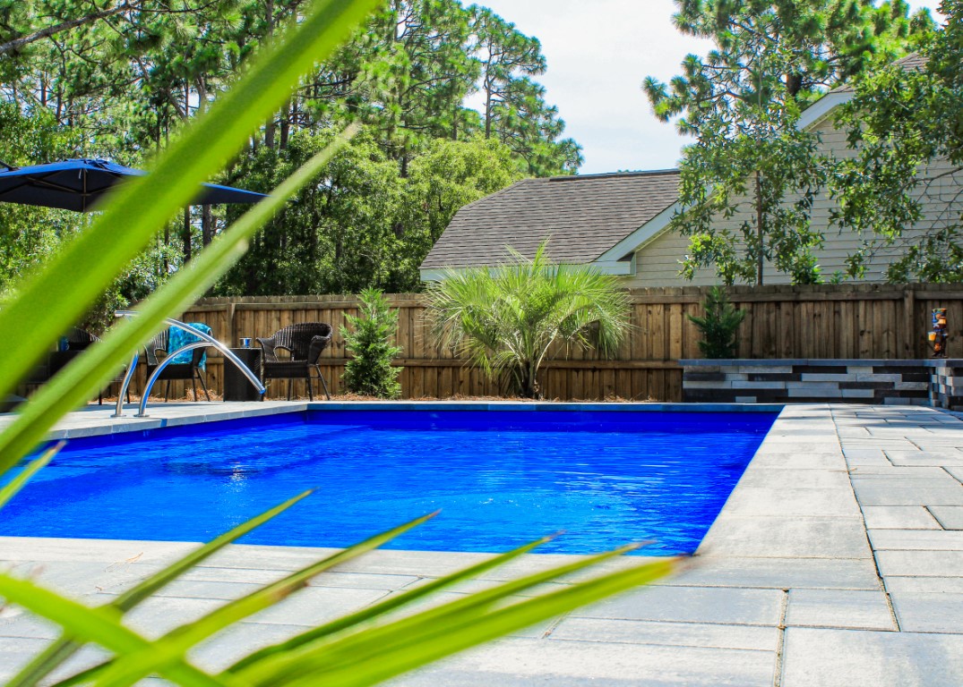 fiberglass inground pool area in residential backyard, concrete surround and fence with green landscaping
