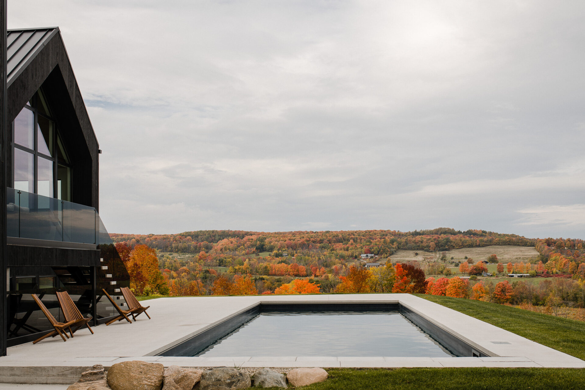 Modern black-clad home with minimalist concrete pool overlooking a vibrant autumn landscape of rolling hills, colorful trees, and open farmland under an overcast sky.