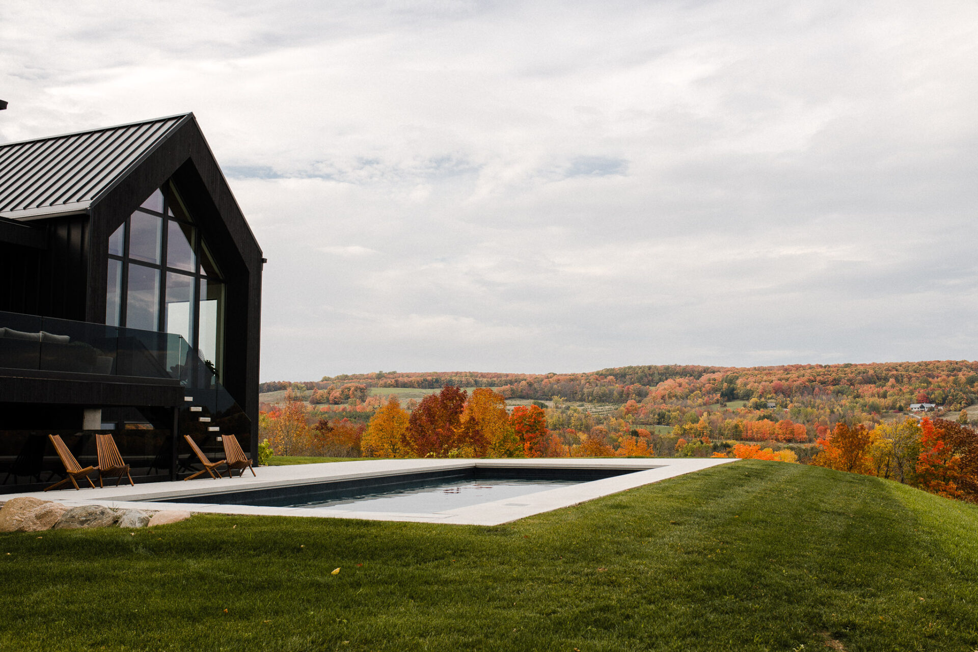 Modern black-clad home with minimalist concrete pool overlooking a vibrant autumn landscape of rolling hills, colorful trees, and open farmland under an overcast sky.