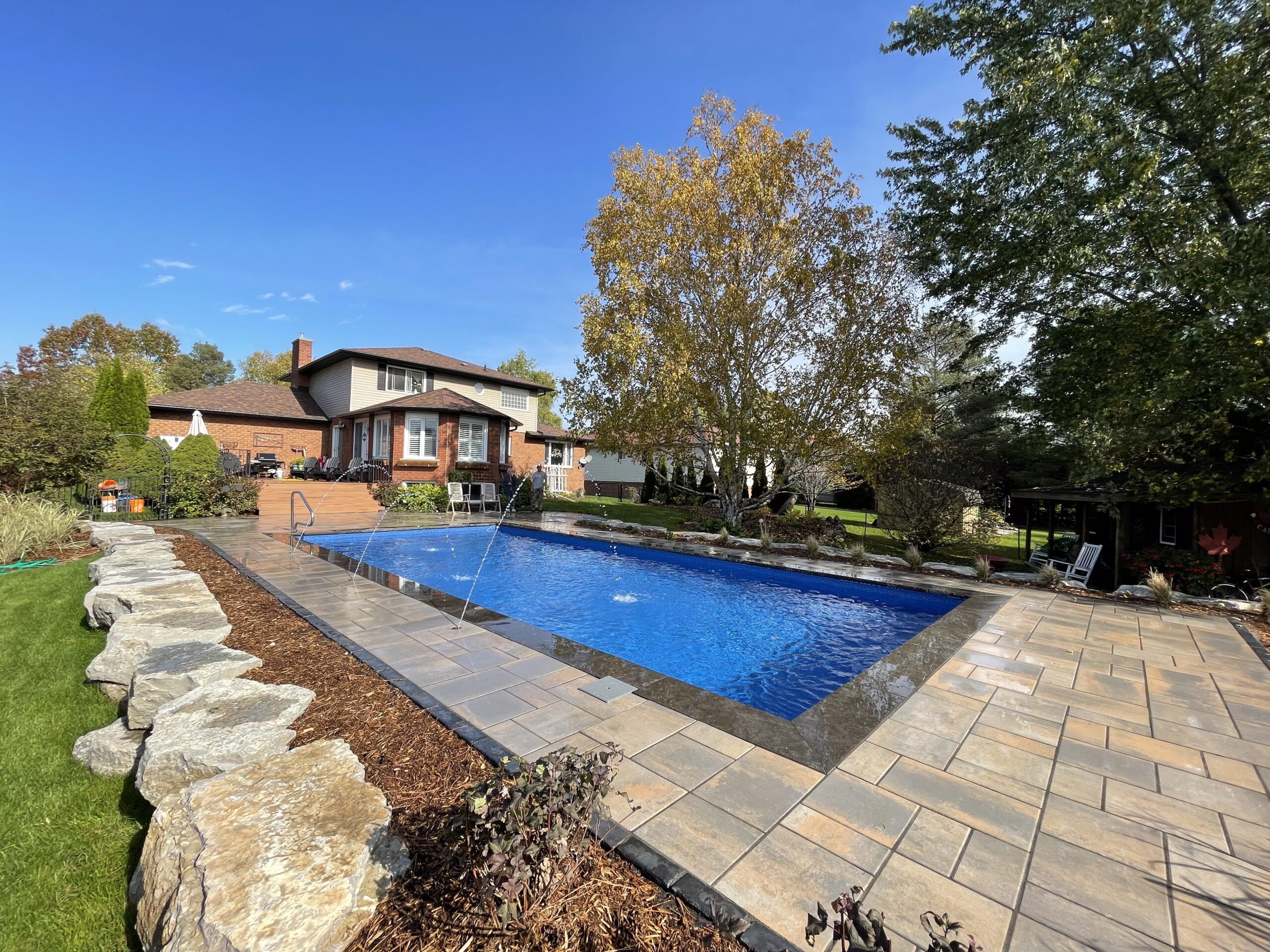 Modern white two-story home with black windows, elevated deck with glass railing, rectangular in-ground pool, stone tile surround, fresh sod, and minimalist landscaping.