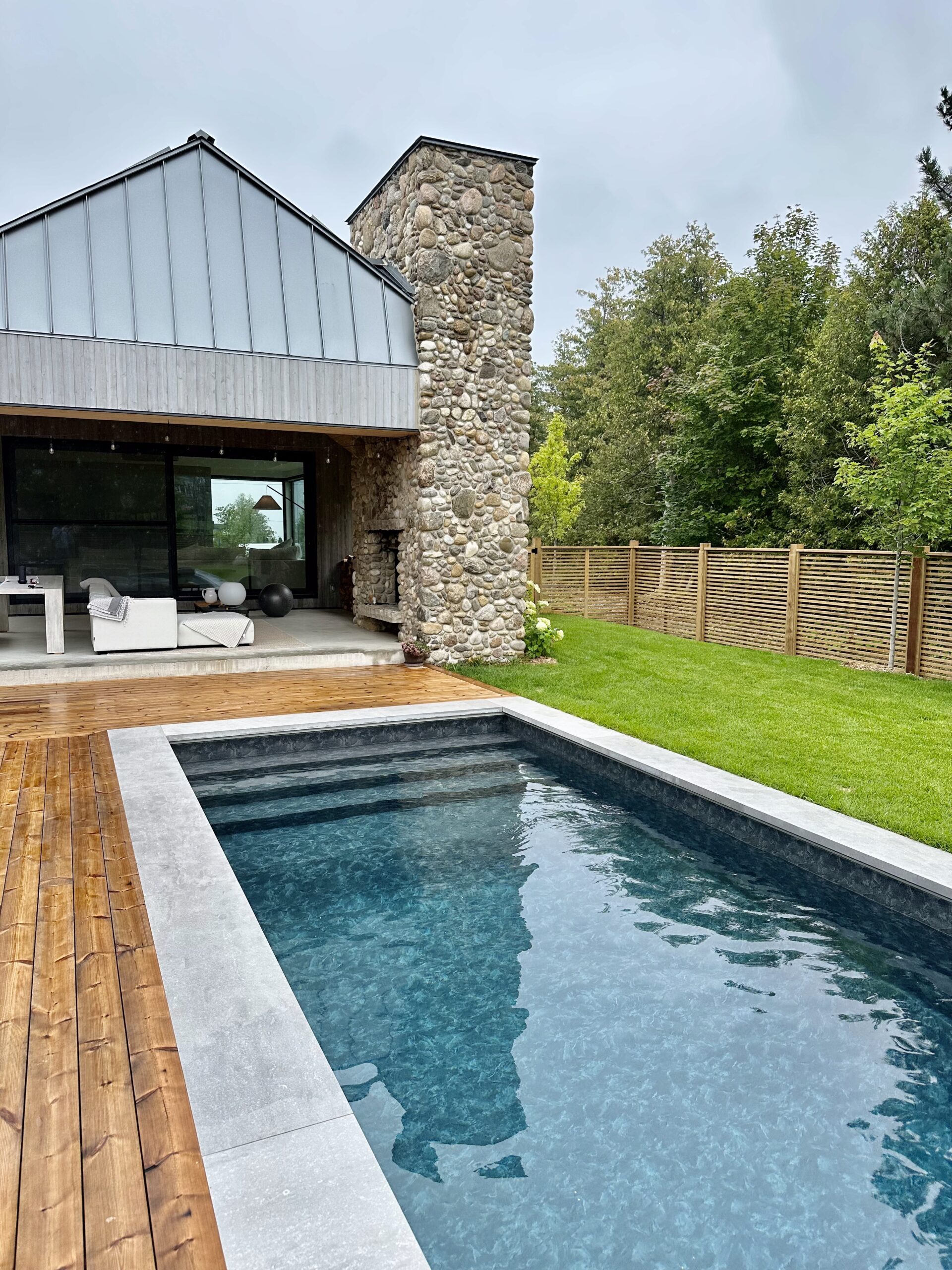 Residential backyard with inground vinyl pool and wooden deck, trees in background and blue sky. Outdoor covered seating area in background, with cobblestone fireplace and modern furniture