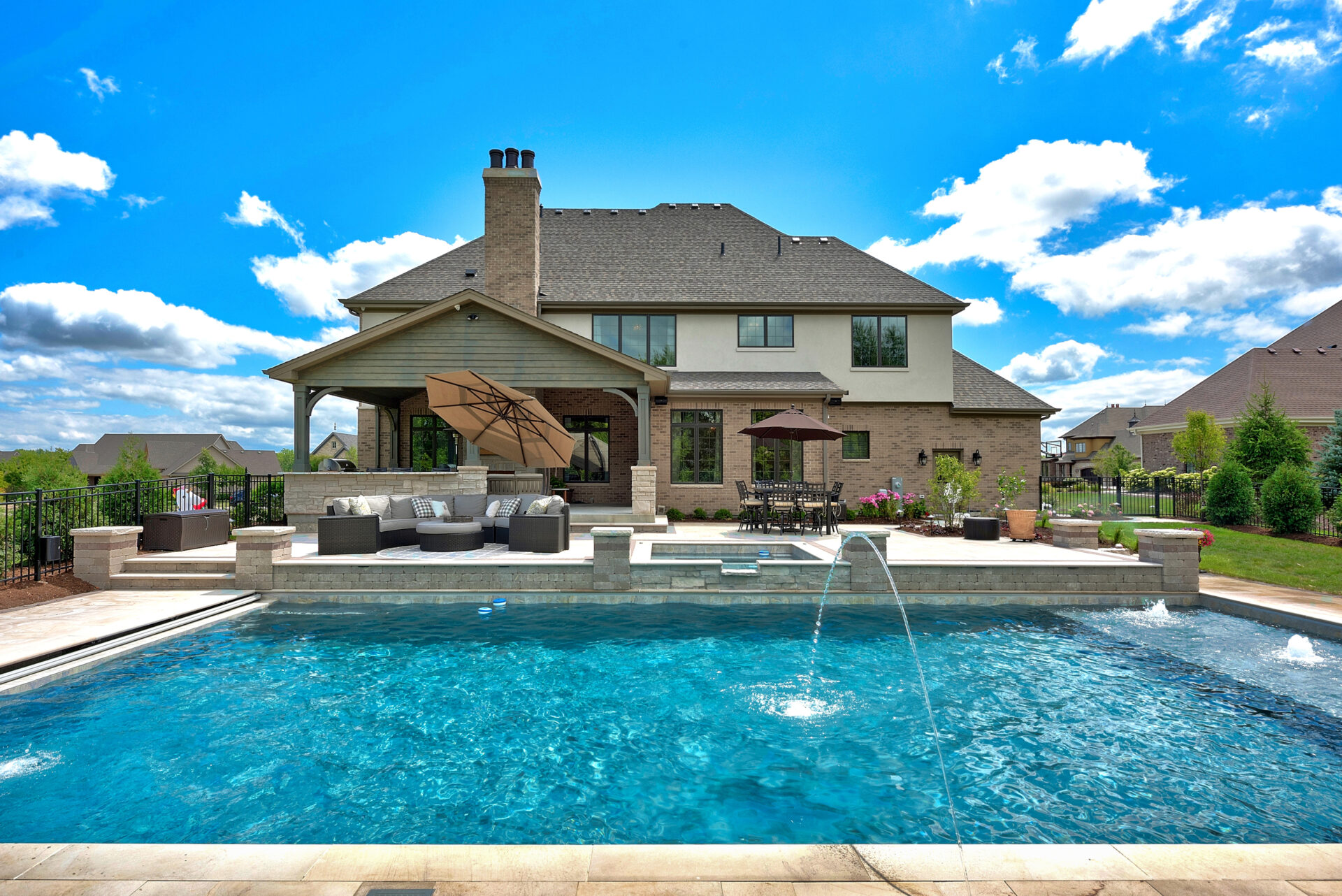 Two-story brick home with backyard pool, elevated lounge area, water features, patio umbrellas, outdoor dining space, and manicured landscaping under a bright blue sky.