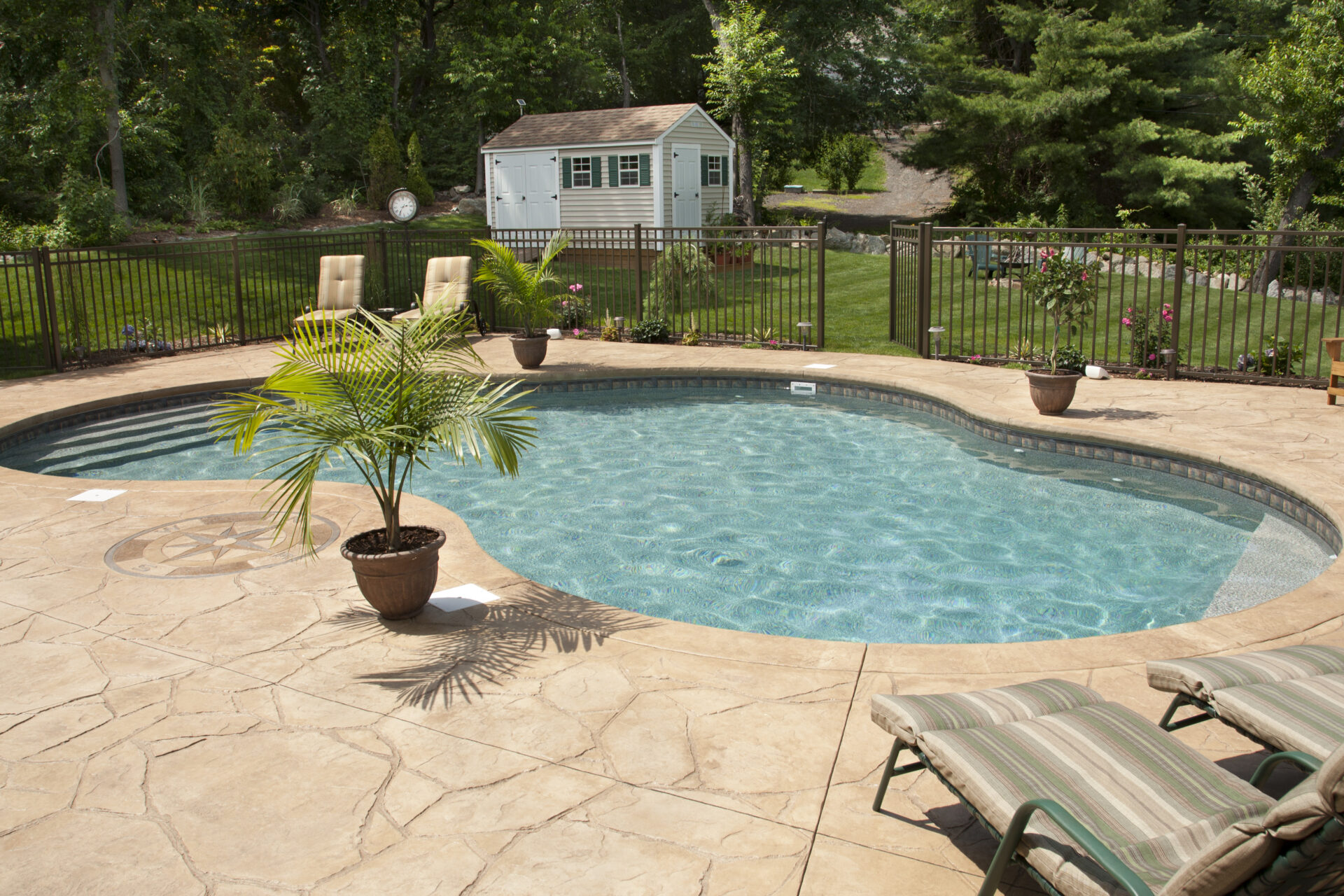 Lush, resort-like backyard salt water swimming pool with flagstone stamped concrete patio. Bronze fencing is seen around pool area.
