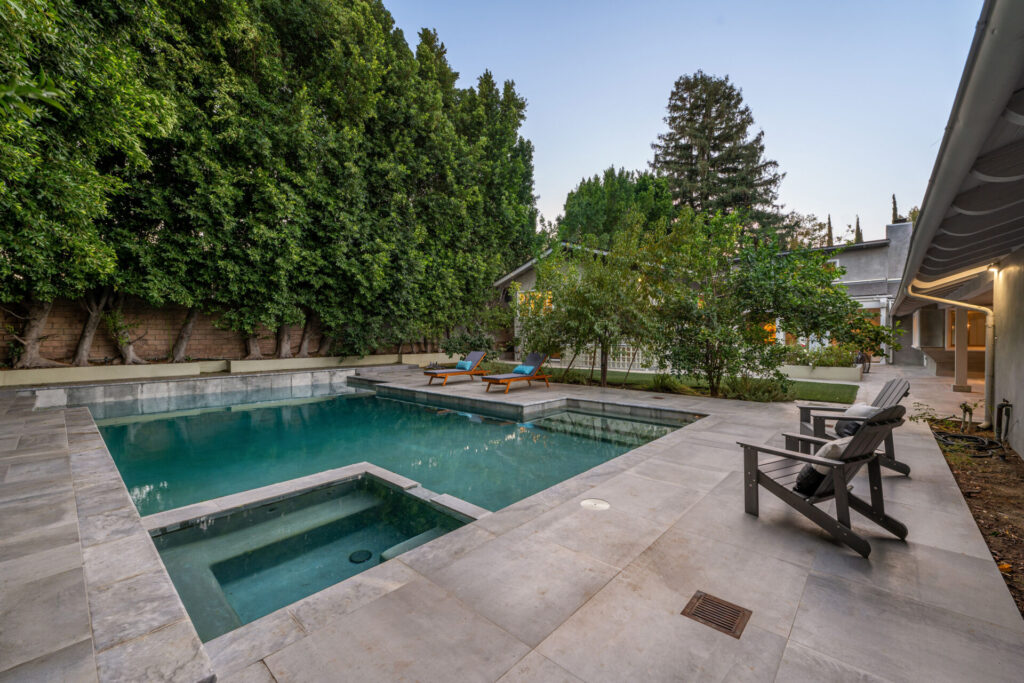 A modern backyard with a swimming pool, lounge chairs, and lush green trees