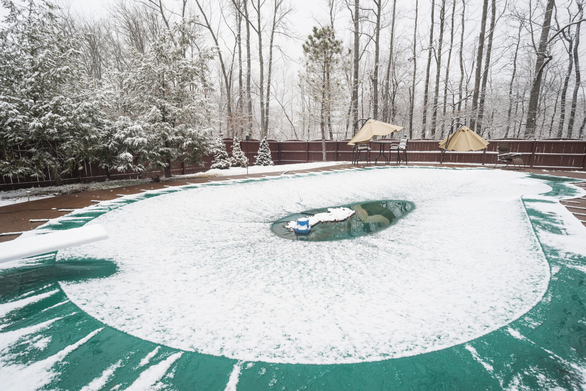 Inground swimming pool with cover on it and water pump during a snowfall in winter
