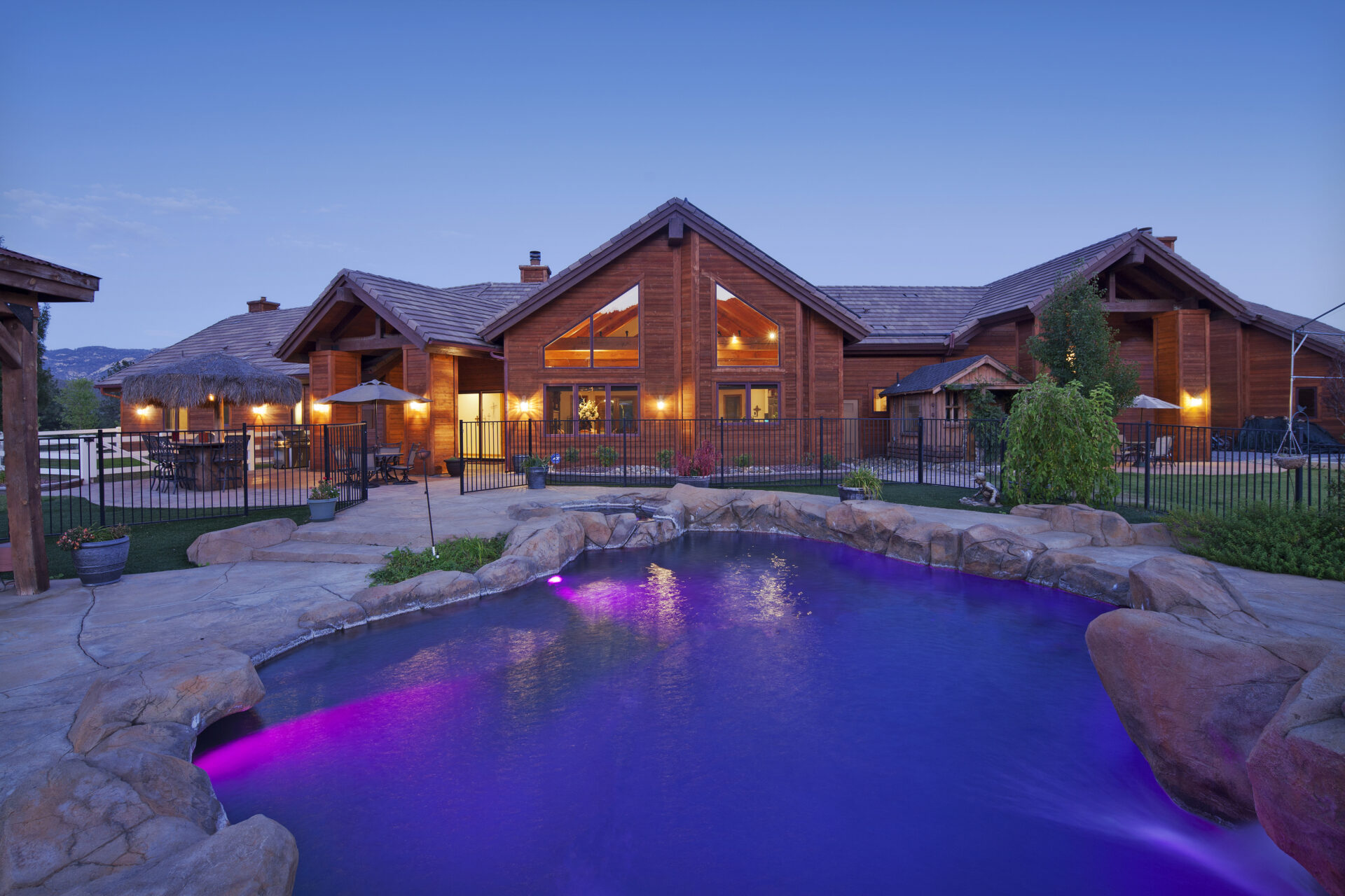 Sprawling log-style ranch home with vaulted gables, large windows, and glowing exterior lights, set behind a freeform rock-edged pool with purple LED lighting and lush landscaping at twilight.