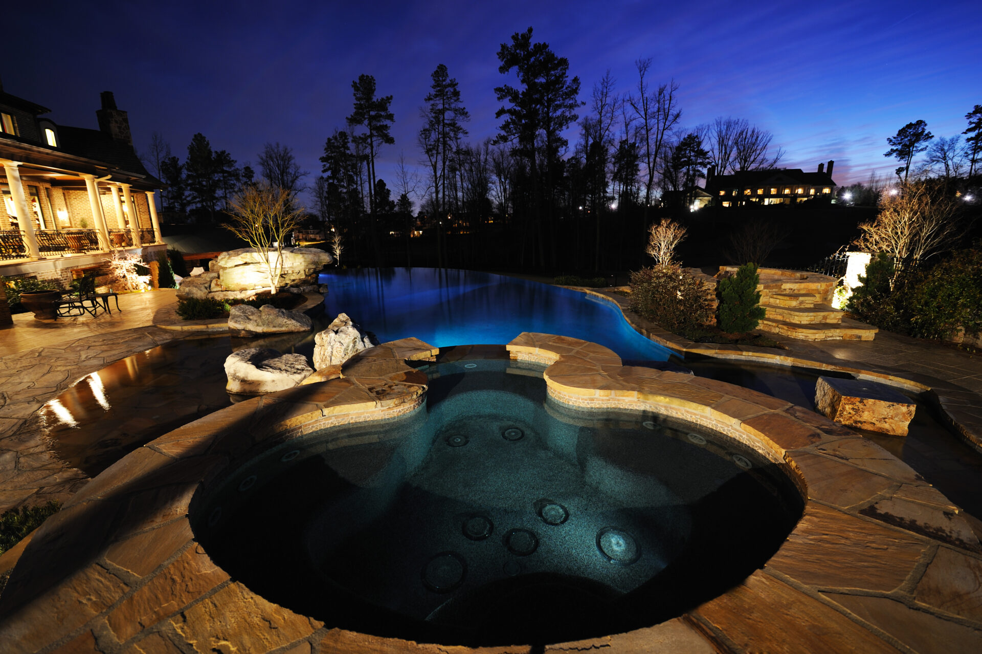 Luxurious backyard with an illuminated infinity pool and attached spa at dusk, surrounded by natural stone decking and landscaping, with a large home and trees in the background.
