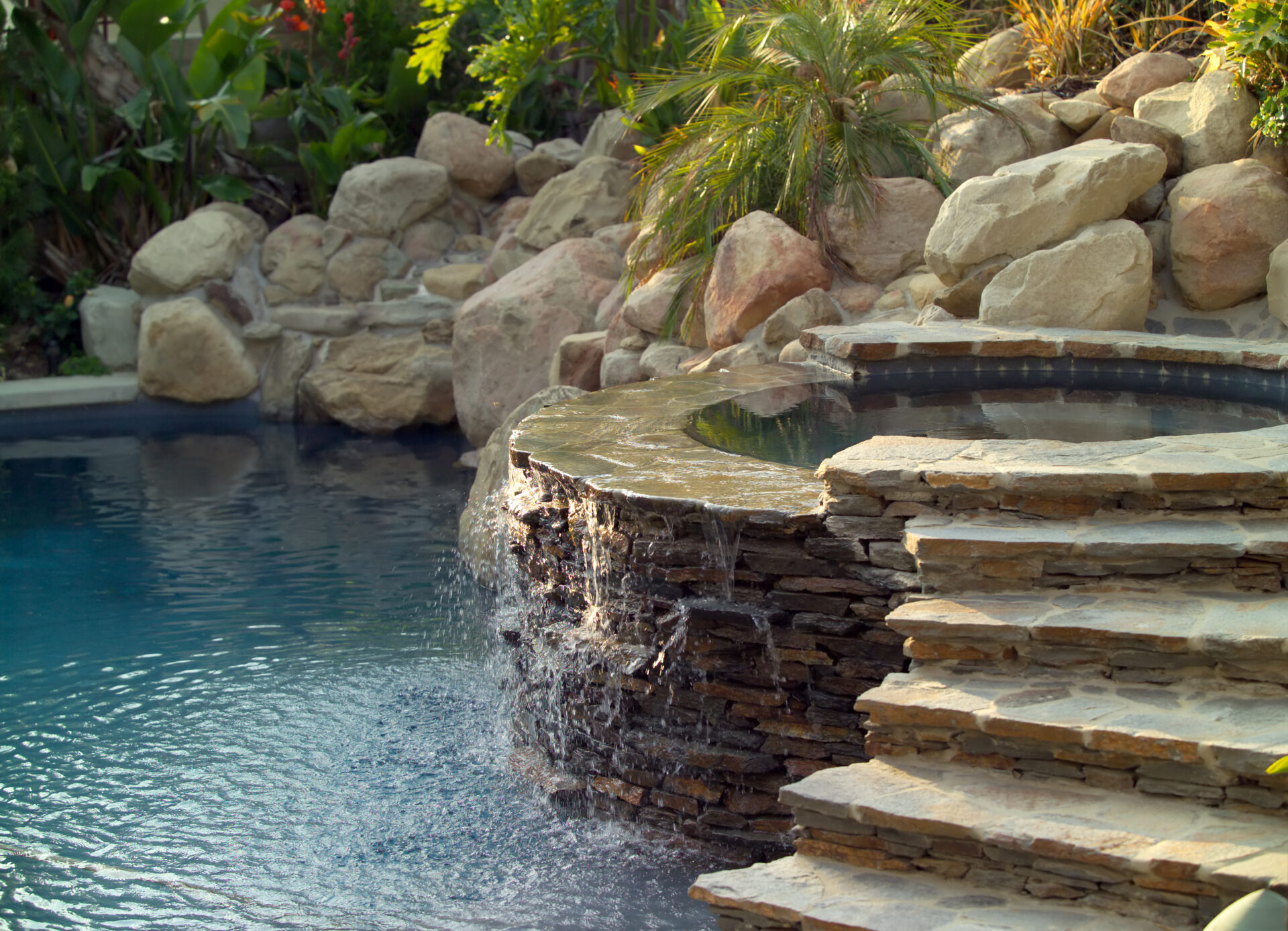 Natural-style backyard pool and spa with a stone waterfall feature cascading from a raised hot tub into the pool below, surrounded by boulders, palm plants, and lush tropical landscaping.