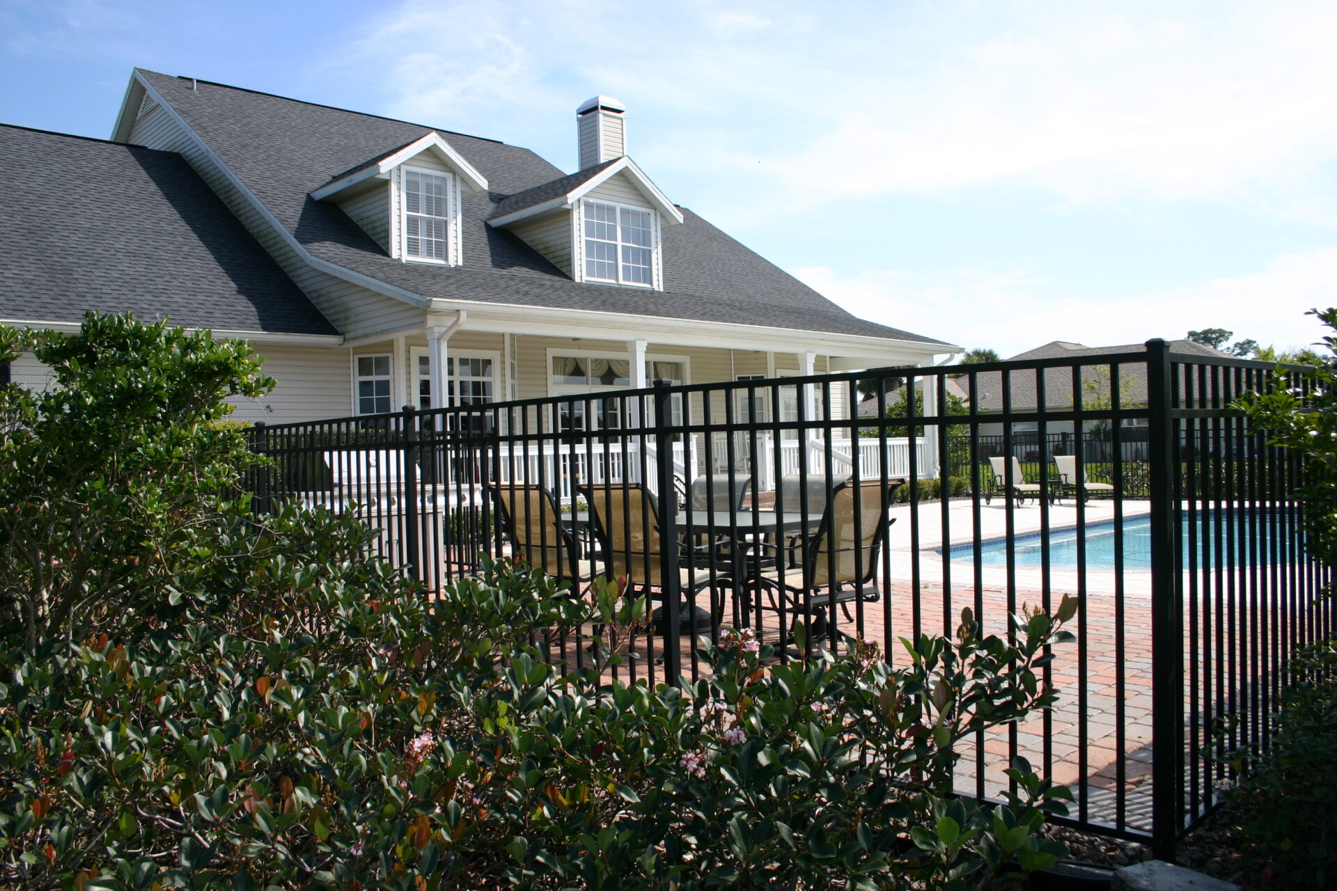 Suburban home with a fenced backyard patio featuring a dining set and an in-ground swimming pool, surrounded by brick pavers and landscaped bushes.