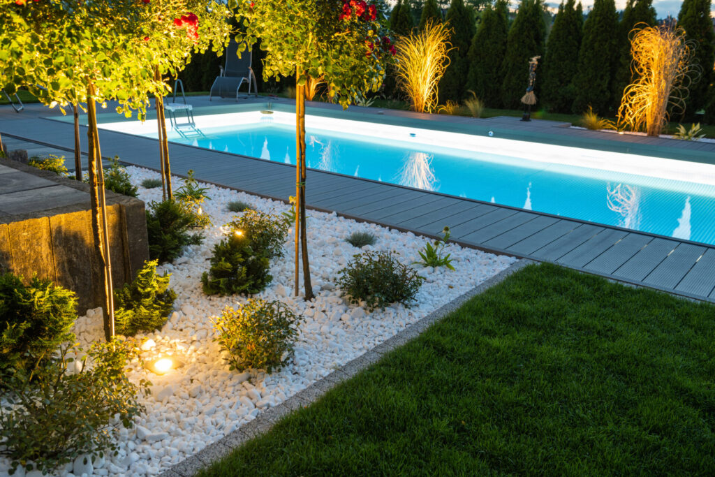 Illuminated modern backyard pool with glowing underwater lighting, surrounded by sleek decking, manicured grass, and a white rock garden with small shrubs and accent trees.