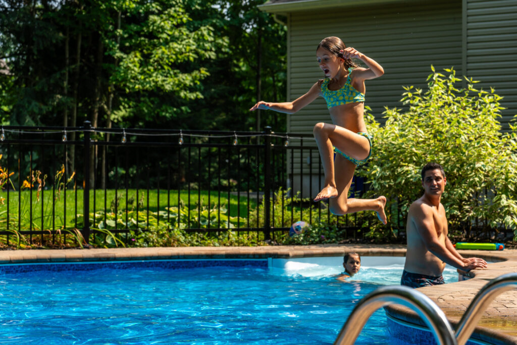A young girl having fun and jumping in her pool in front of her father and sister during a sunny day of summer.