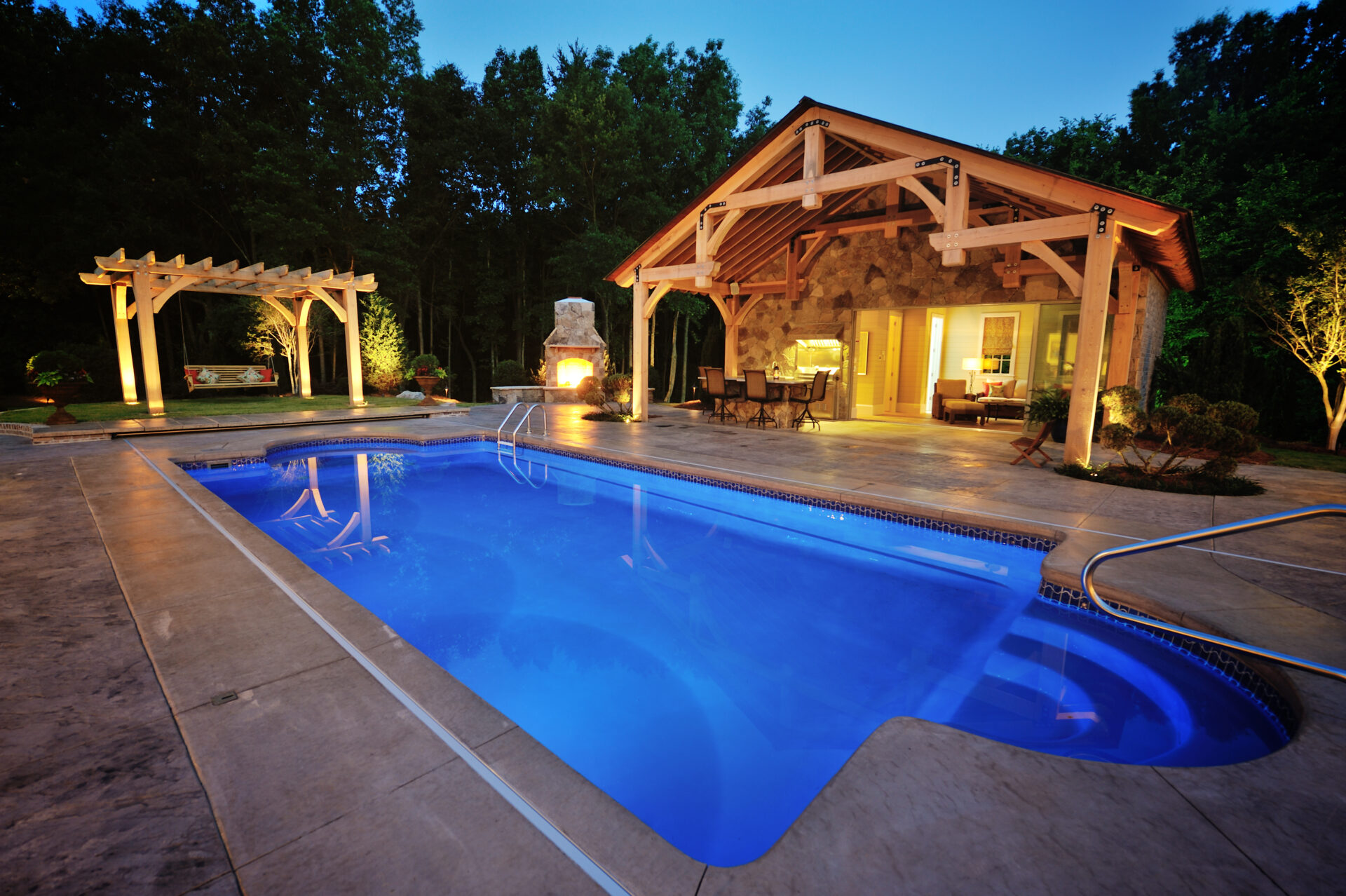 Luxury backyard with a glowing in-ground pool, stone patio, pergola swing, and a rustic pool house featuring an outdoor fireplace, all softly lit against a wooded backdrop at dusk.
