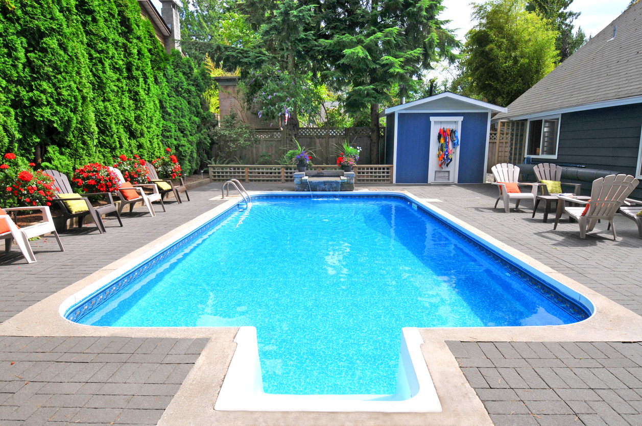 Backyard inground pool surrounded by lounge chairs with colorful cushions, vibrant red flowers, and a small blue pool house, set against a backdrop of tall evergreen trees.