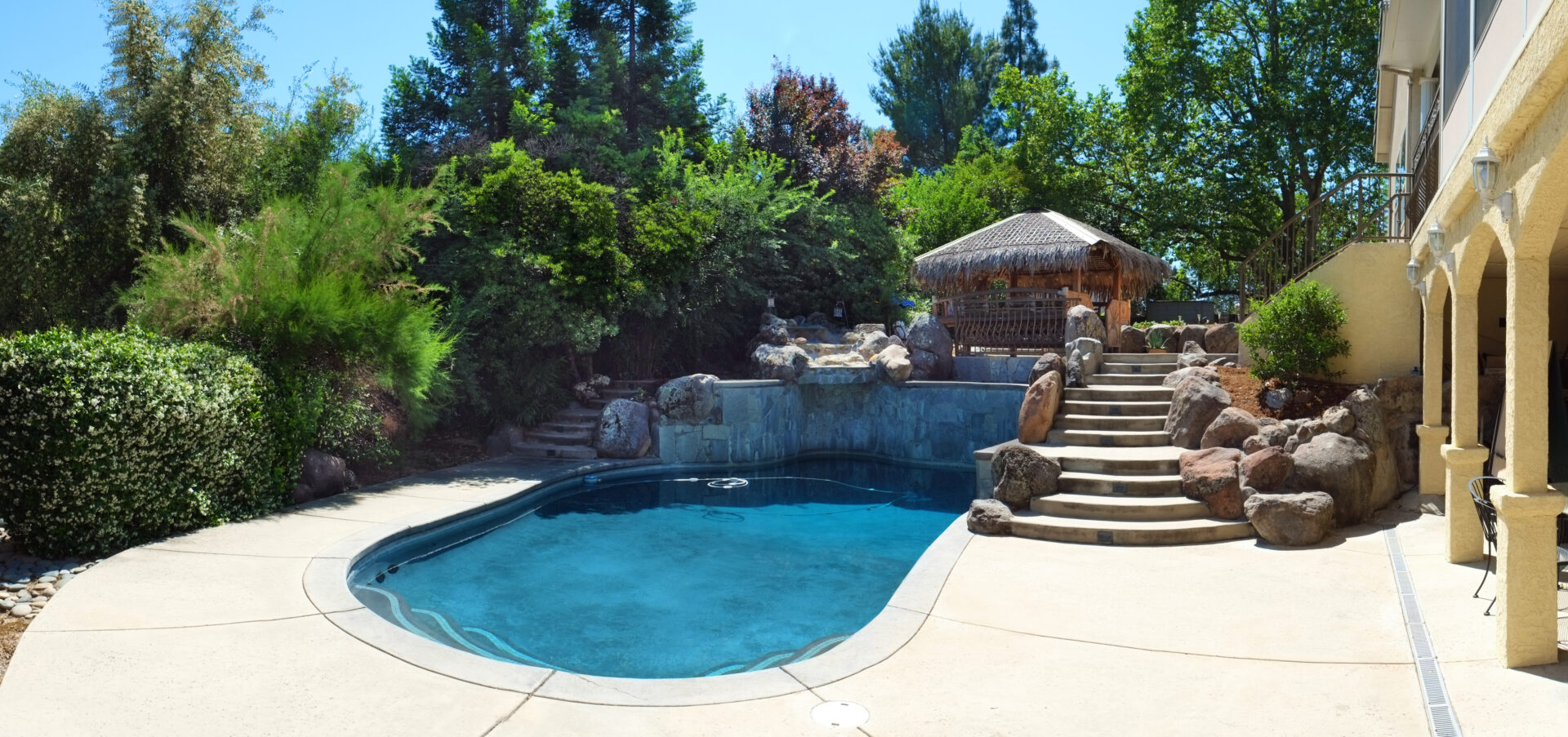 Backyard pool surrounded by lush greenery and natural stone landscaping, with curved steps leading to a thatched-roof tiki hut seating area and tropical garden elements.