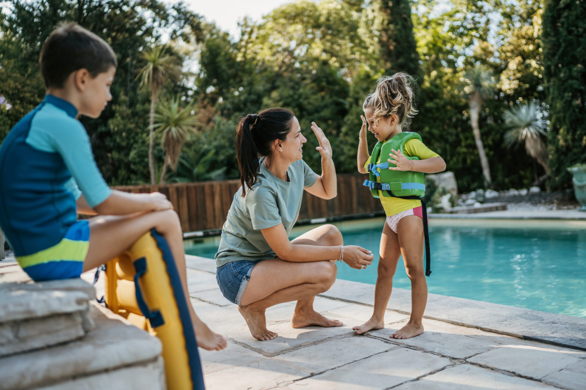 Mother giving a high-five to her young daughter in a green life jacket by the poolside, while a young boy in a swim outfit sits nearby holding a pool float.