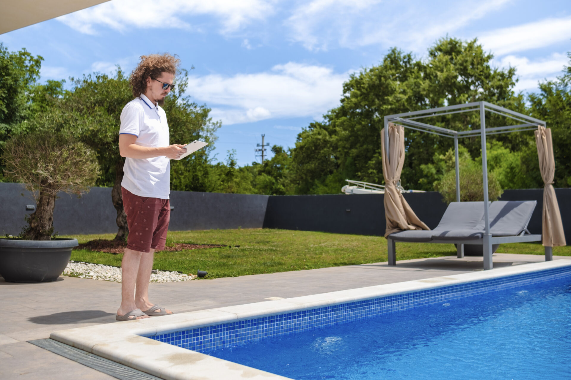 Man in sunglasses and summer clothes standing poolside with a clipboard, inspecting or checking something near a modern residential swimming pool.