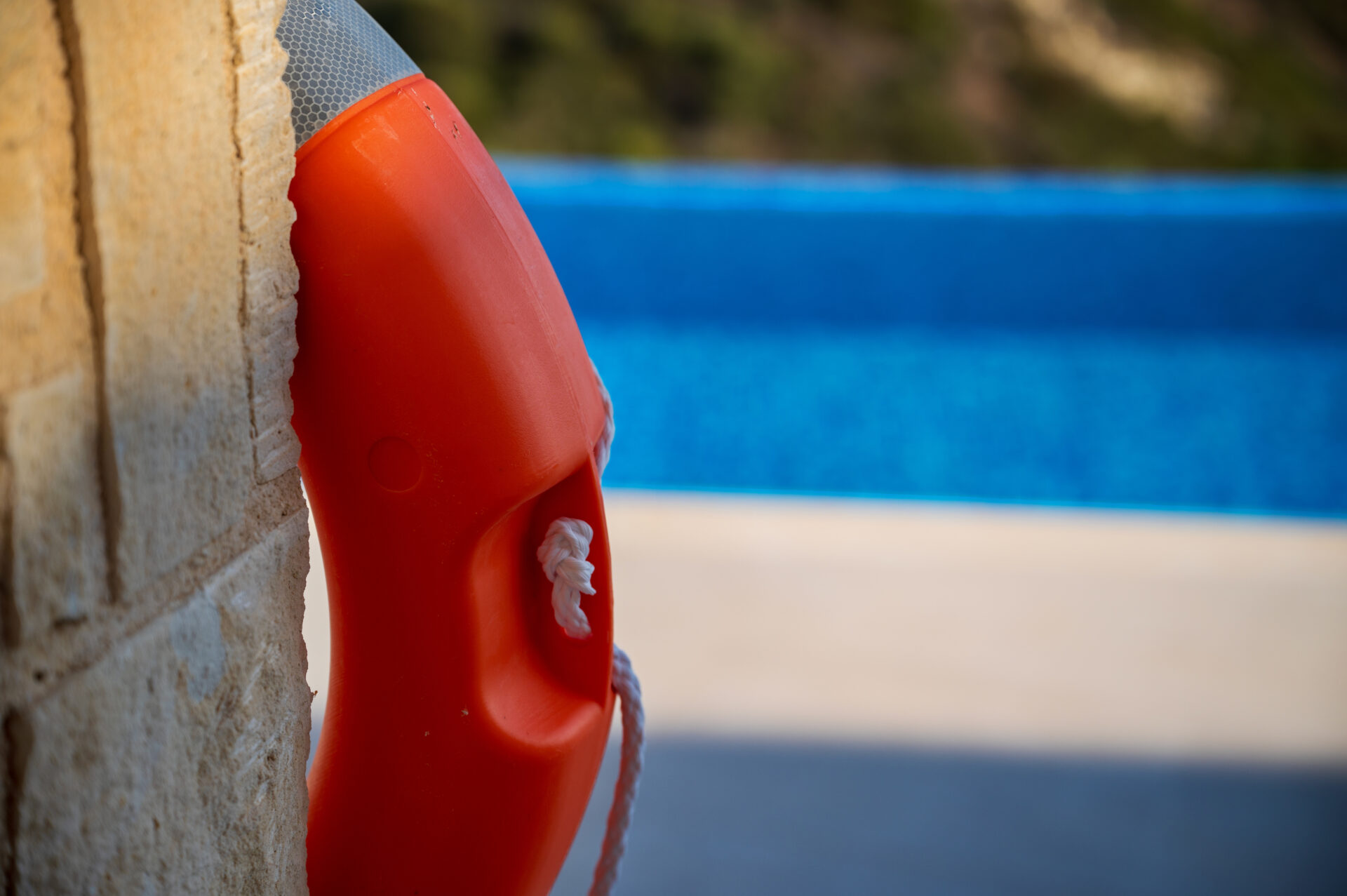Close-up of an orange life preserver with a white rope hanging on a stone wall near a swimming pool, with the blue water blurred in the background.