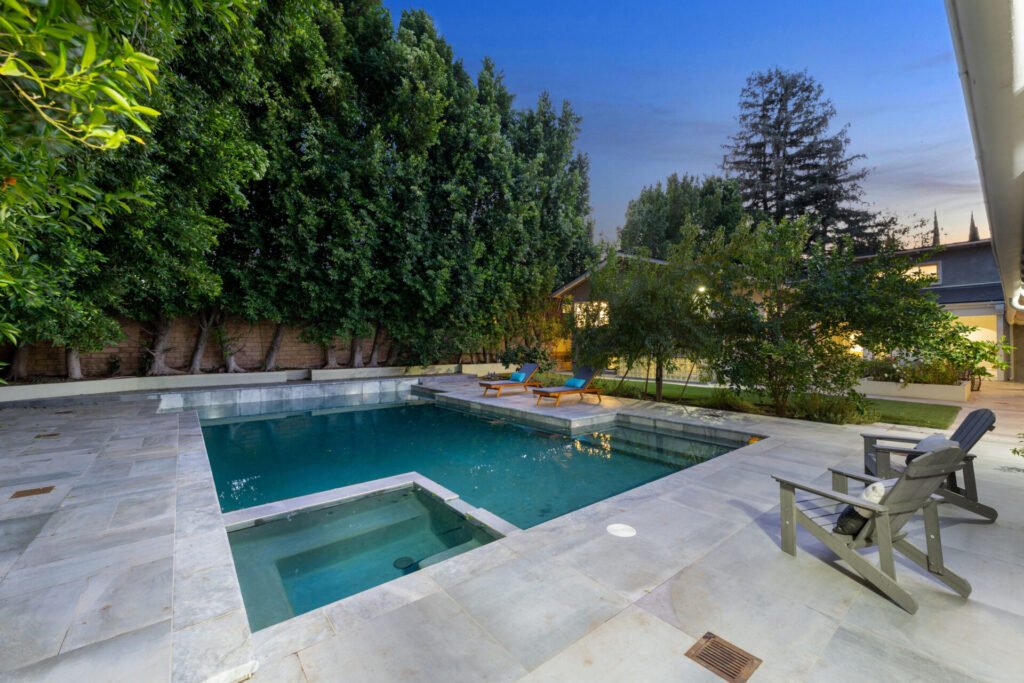 A modern backyard with a swimming pool, lounge chairs, and lush green trees