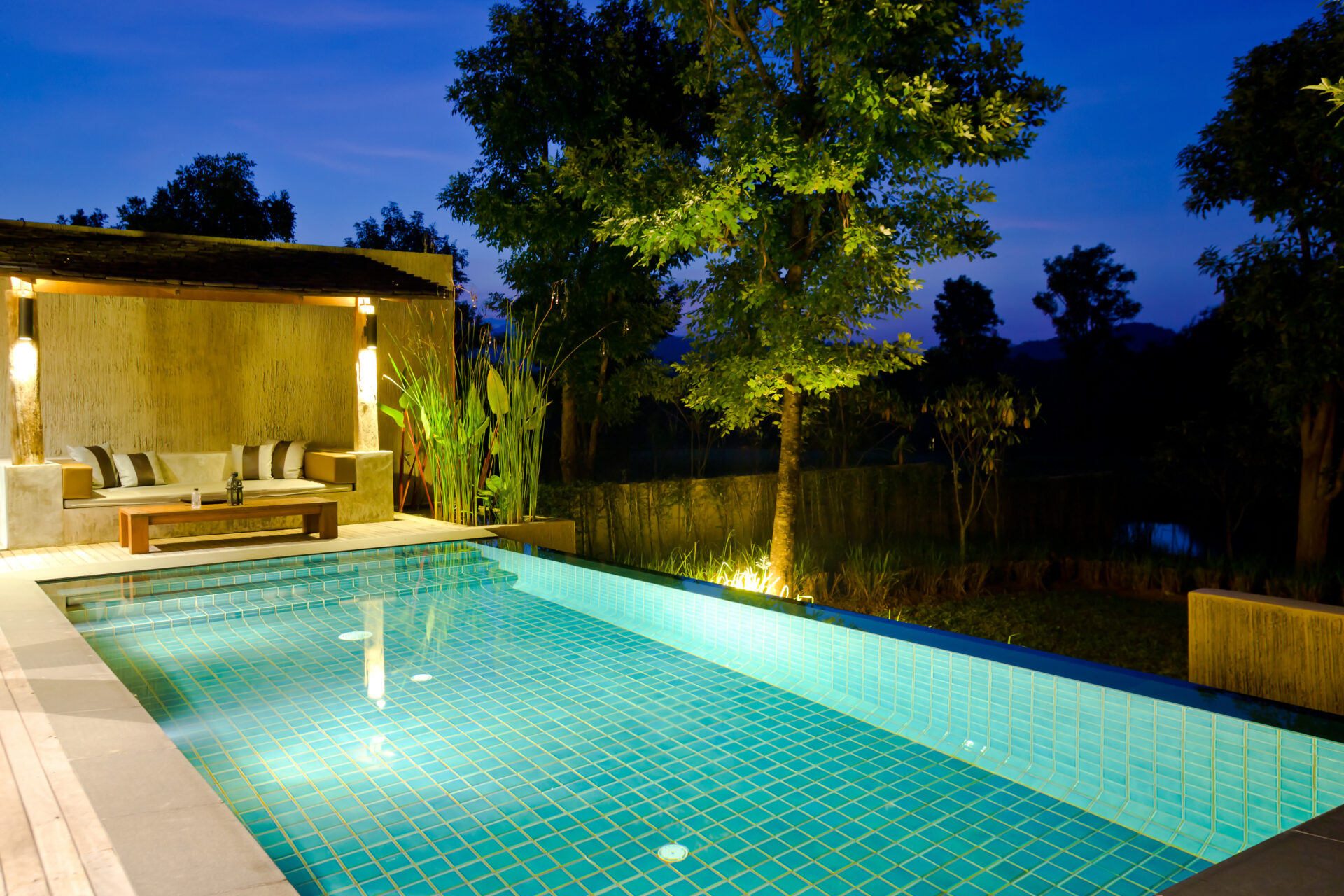 Infinity-edge pool with turquoise tile glowing at dusk, beside a modern outdoor lounge area with a built-in concrete sofa, surrounded by tropical greenery and ambient lighting.