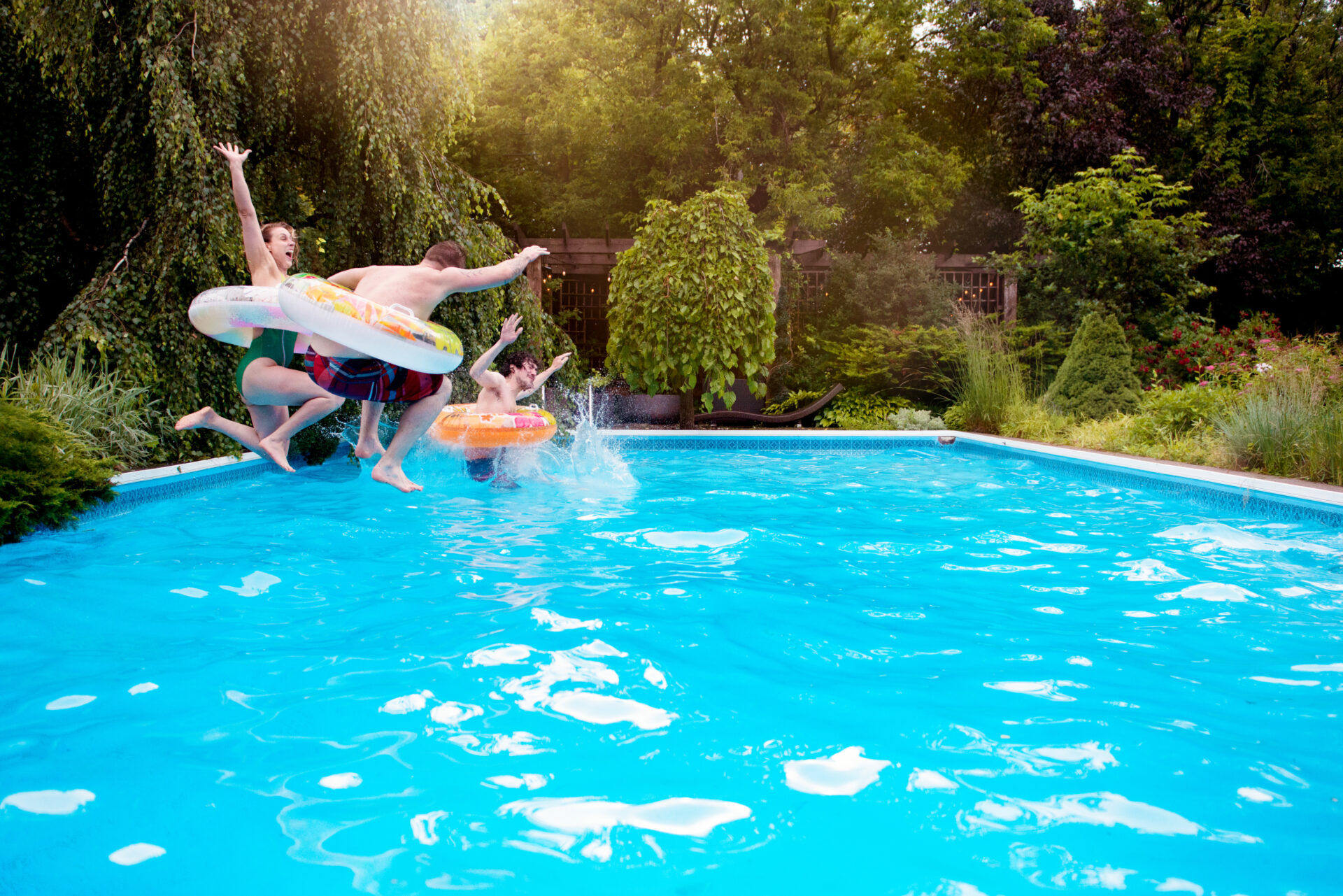 Group of teenagers jumping into a backyard pool with inflatable rings, surrounded by lush greenery and summer foliage.